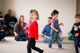 Preschoolers dancing in a circle, smiling and singing, while parents tap and clap to the beat