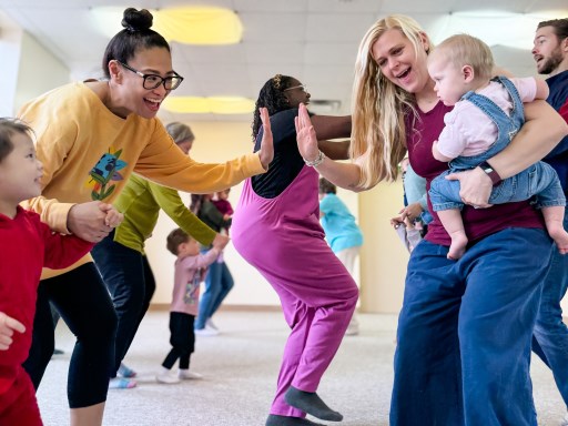 Mom holding preschooler's hand and giving a high five to another mom holding a baby as they enjoy a line dance in music class