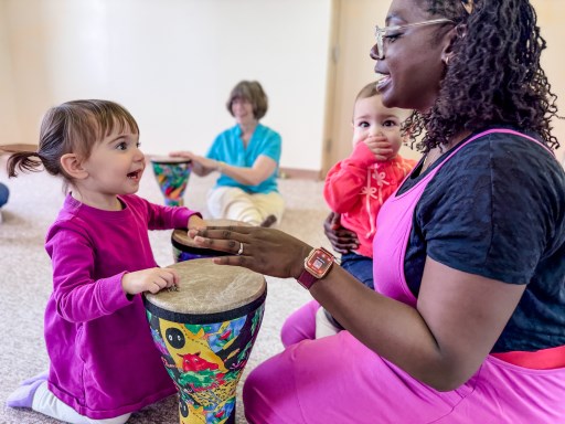Early childhood music teacher is drumming with a happy preschooler and a delighted baby in her arms