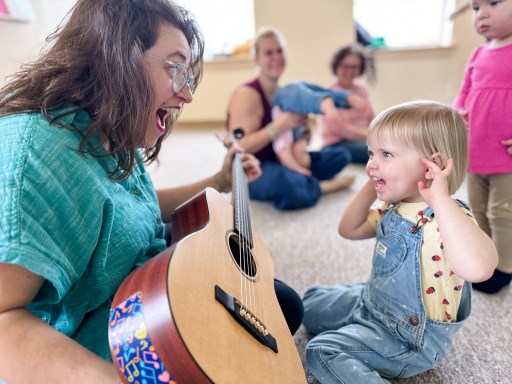 Music teacher joyfully showing her guitar to a preschooler who is delighted to strum & listen to the sound of the instrument