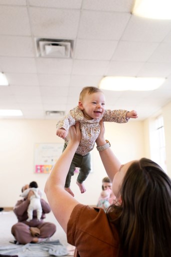 Babies all gathered around a large drum in music class with their moms sitting behind them for support
