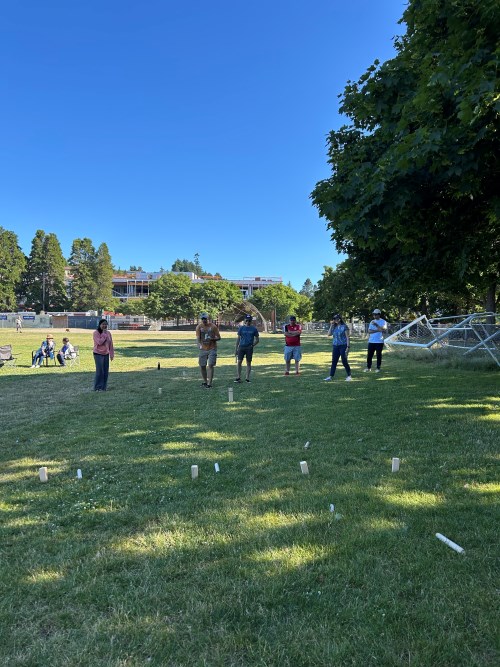Participants playing Kubb on the grass, with wooden game pieces spread out across the field and trees providing shade in the background.