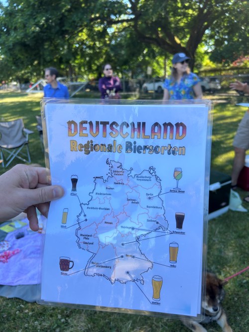 A hand holding a laminated map of Germany showing different regional beer types, with people chatting and relaxing in the background at a park picnic.